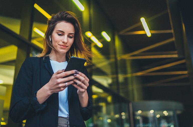Successful female banker using smartphone outdoors while standing near his office background yellow neon lights, young woman professional manager working on mobile device near skyscraper at night